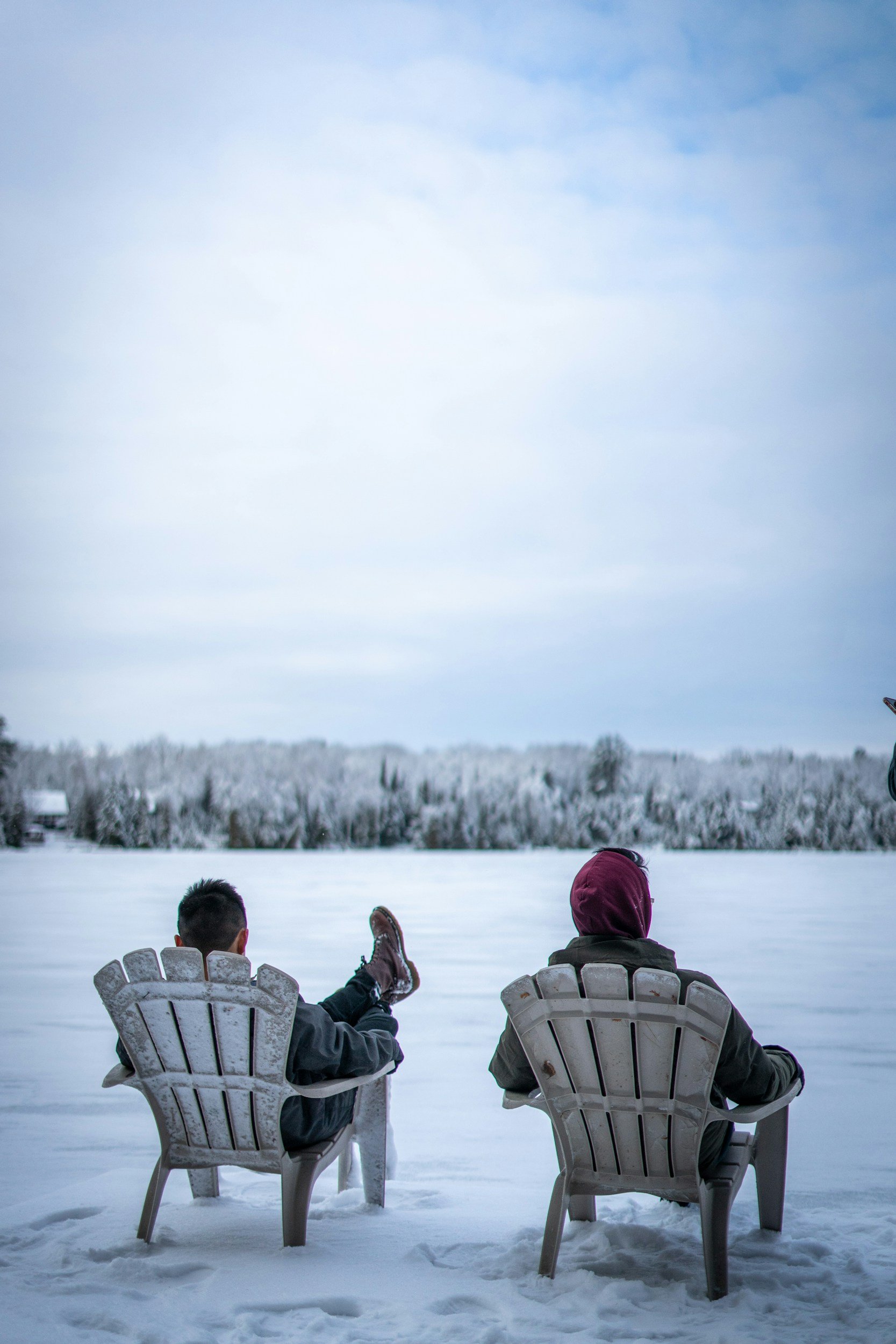 Two people seated in white chairs on a snow-covered lake, looking out towards a distant tree line under a pale blue sky.