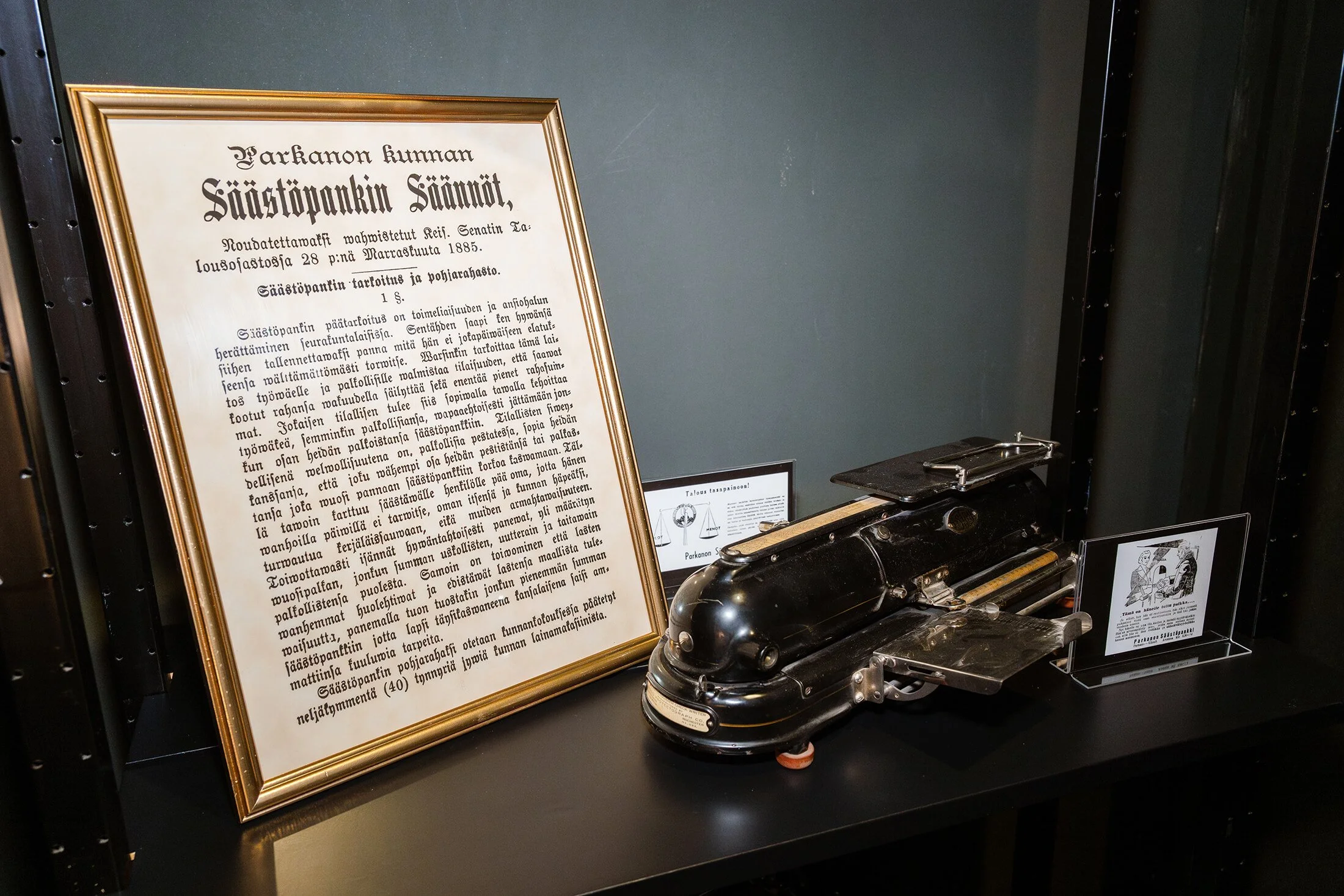 Valokuva museoesineistä, joissa on vanha puhelin ja kehys, jonka sisällä teksti suomeksi.
