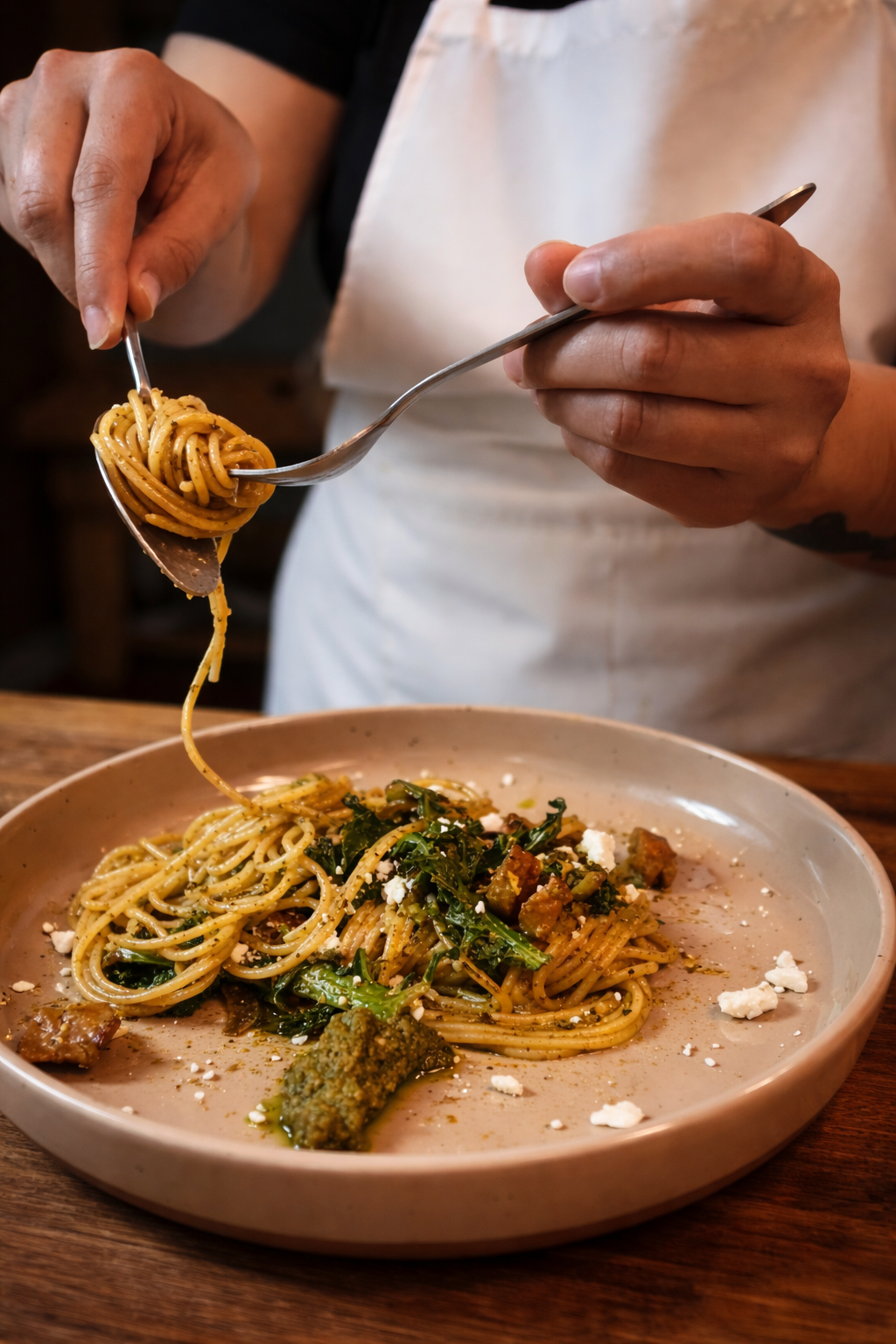 Person in apron eating spaghetti with vegetables and cheese on a beige plate.