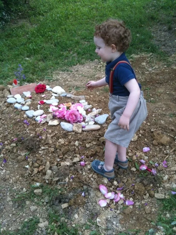 Un jeune garçon avec des cheveux roux, portant une chemise bleue, un short gris et des sandales, se tenant devant une petite tombe décorée de pierres, de fleurs en papier, de pétales de fleurs et de petites fleurs violettes sur le sol en terre.