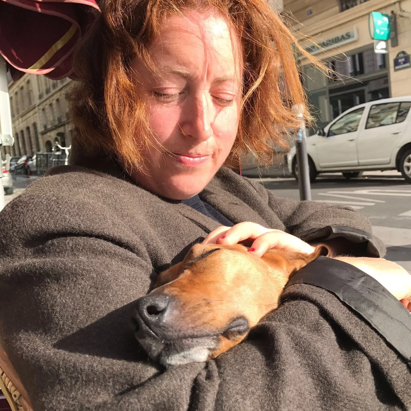 Une femme avec des cheveux bouclés rouges tient un chien couché dans ses bras, affichant une expression paisible et heureuse, dans une rue urbaine ensoleillée.
