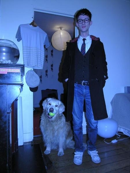 Jeune homme avec des lunettes, portant un manteau noir et une cravate, debout à côté d'un chien golden retriever avec un tennis dans la bouche à l'intérieur d'une pièce domestique.