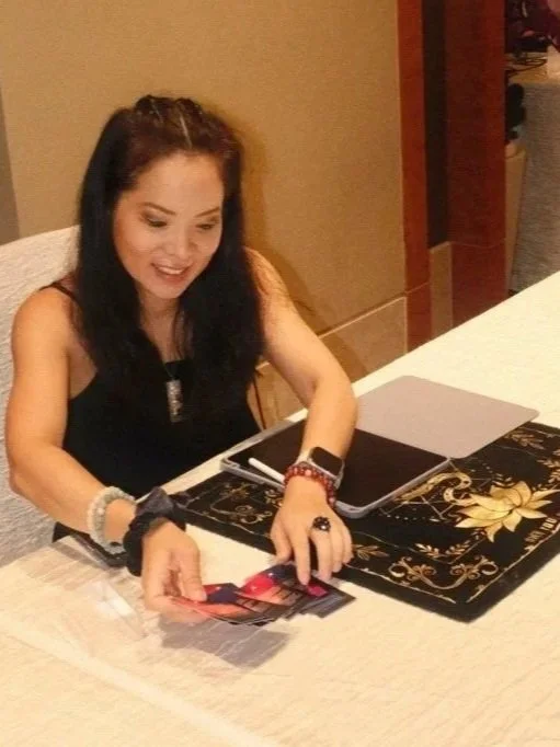 A young woman with dark hair sitting at a table, smiling, holding a photograph. The table has a black and gold decorative runner and items including a closed laptop and a smartphone.