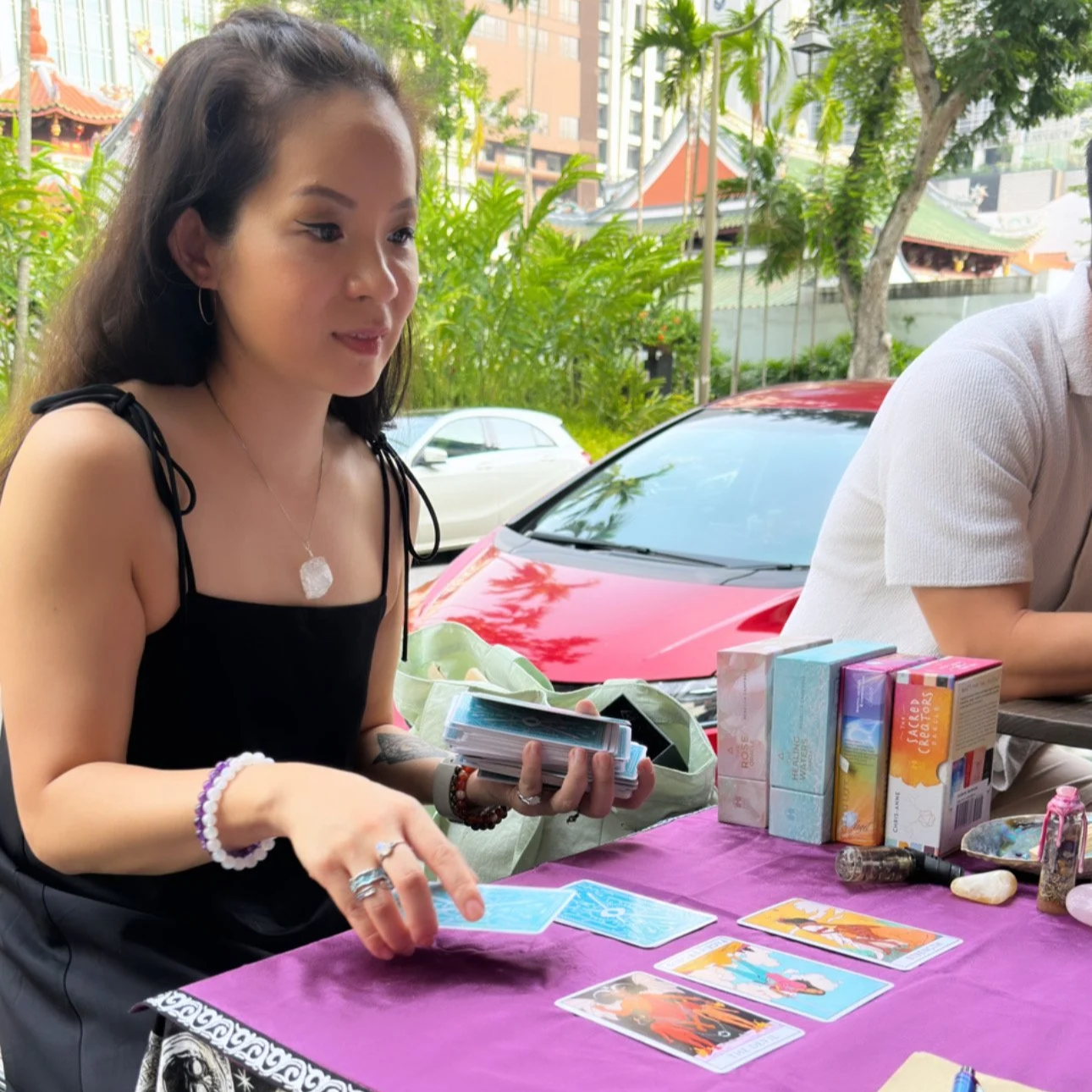 A woman with long dark hair, wearing a black tank top and jewelry, is sitting at a table with tarot cards laid out in front of her, with boxes of tarot decks and crystals nearby. She is outdoors with trees, a red car, and buildings in the background.
