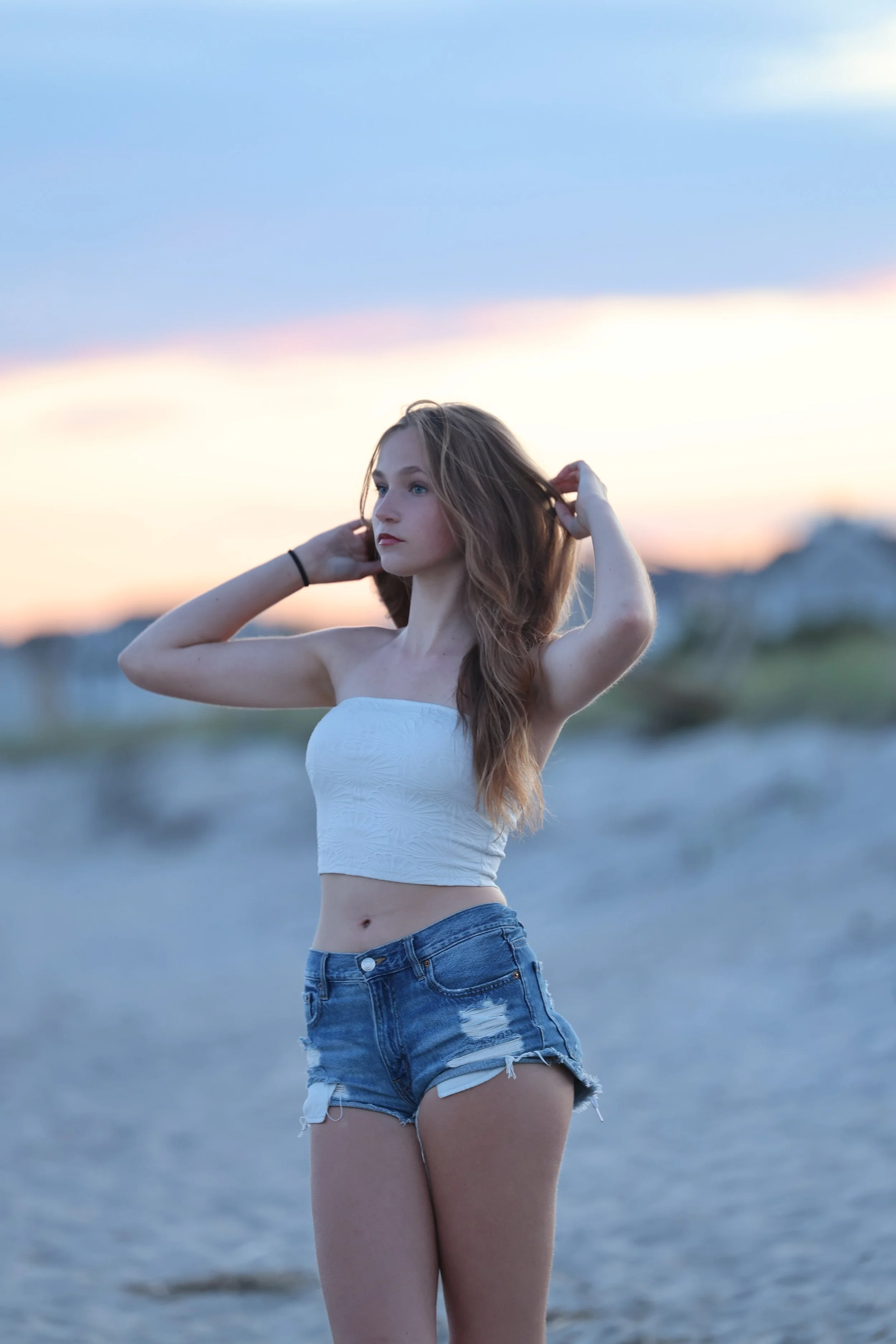 A young woman with long, wavy hair standing on a beach during sunset, wearing a white strapless top and denim shorts.