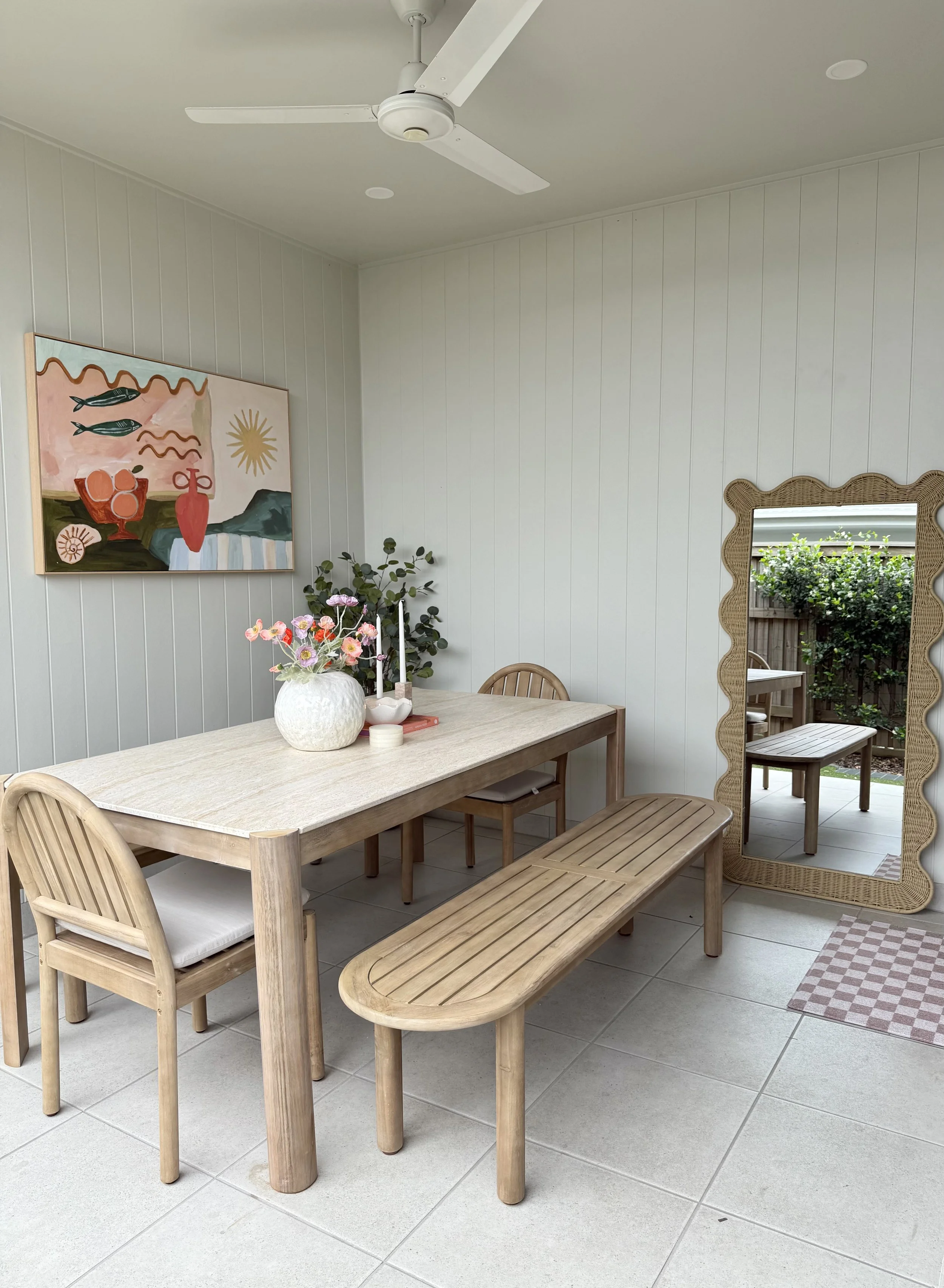 A dining area with a light wood table, a bench, and three chairs. The table has a white vase with pink and purple flowers, candles, and a small dish. A large, ornate mirror leans against the wall, reflecting part of a garden. A framed abstract art piece hangs on the wall, and a ceiling fan is above the table.