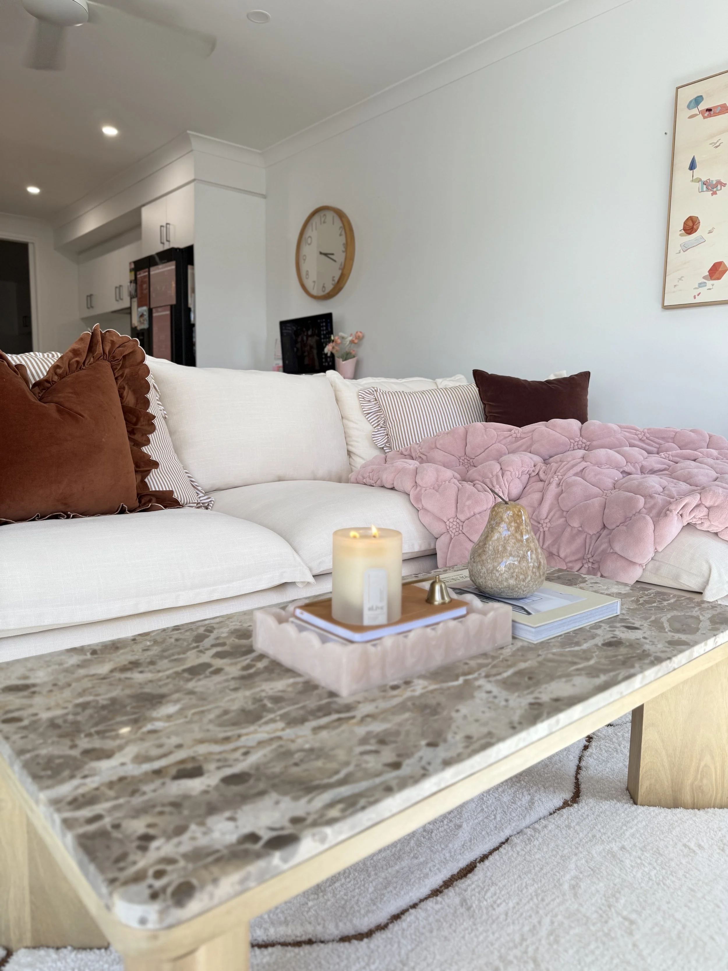 A living room with a white sofa decorated with brown, striped, and pink pillows. A pink plush blanket is on the sofa. In front of the sofa is a marble coffee table with a candle, decorative rock, and books. The background shows a wall clock, a small TV, and artwork on white walls.