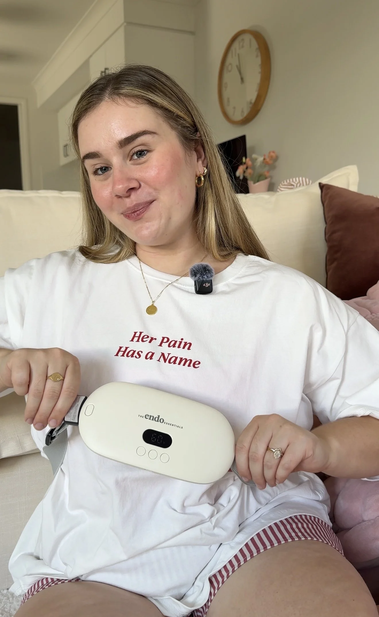 A woman sitting on a couch holding a device with a digital screen and buttons, wearing a white t-shirt with red text that reads 'Her Pain Has a Name', showing a slight smile, with a clock and flower vase in the background.
