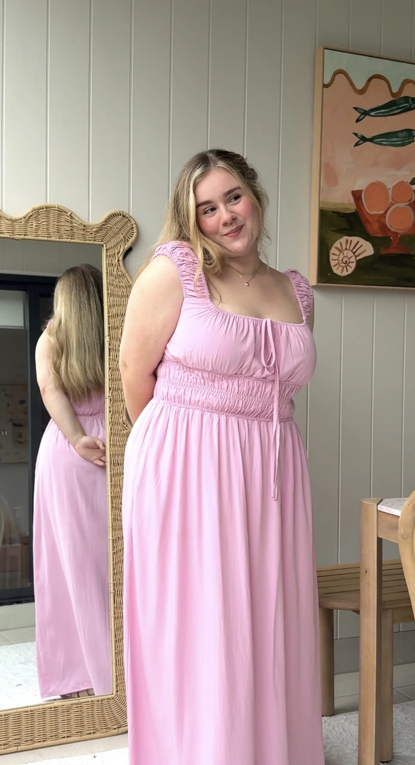 A young woman with blonde hair wearing a pink fayt the label dress standing in front of a mirror, with a colorful painting on the wall behind her.
