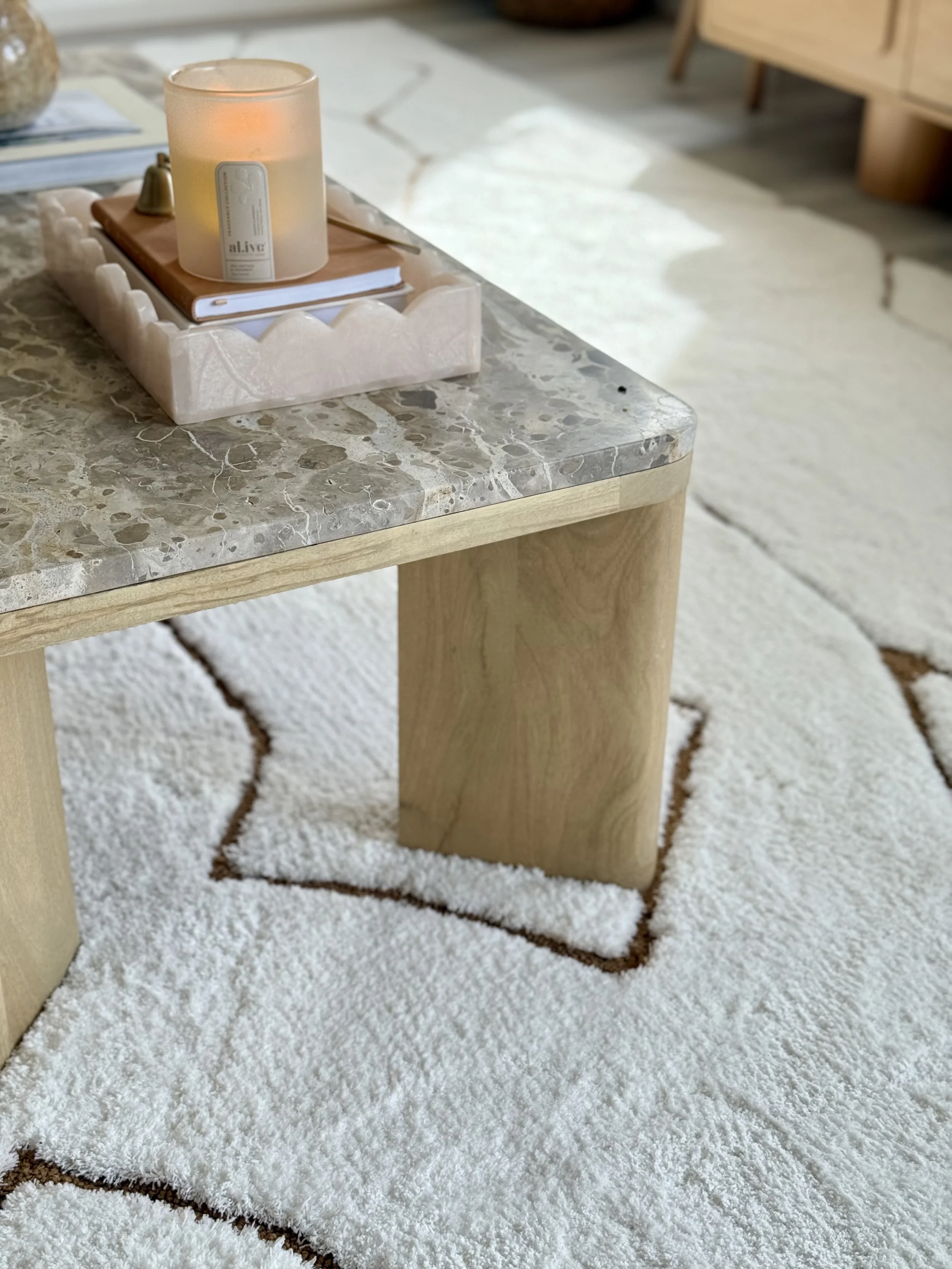 Close-up of a marble-topped wooden coffee table with a pink tray holding a candle, notebook, and a small bell, placed on a textured white rug with brown lines.