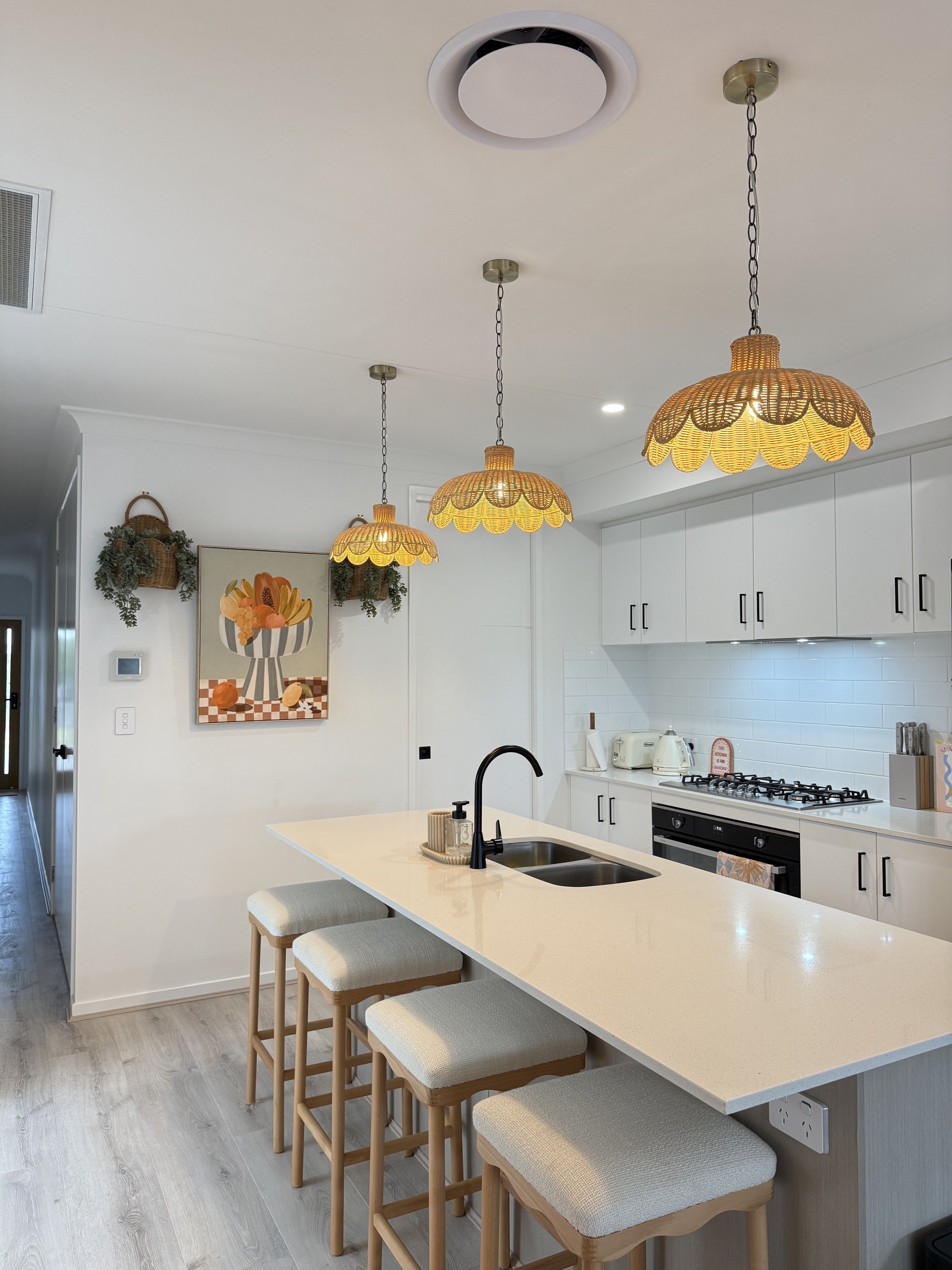 Modern kitchen with white cabinets, a large white island with four beige upholstered stools, a black sink faucet, and three decorative pendant lights with yellow scalloped shades hanging from the ceiling. There is a painting of a vase with flowers on the wall, and small kitchen appliances on the counter.