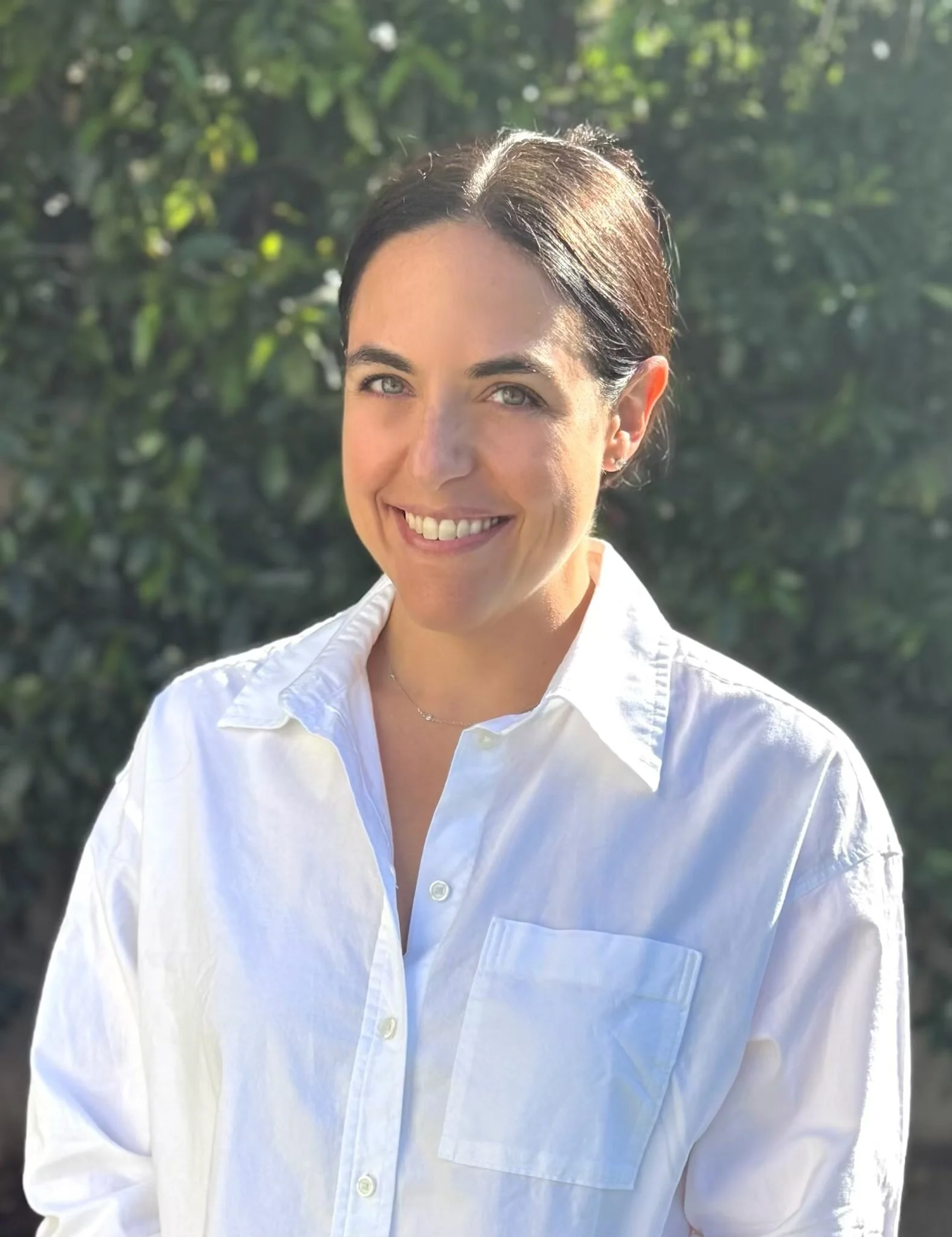 A smiling woman with dark hair, wearing a white button-up shirt, standing outdoors with greenery in the background.