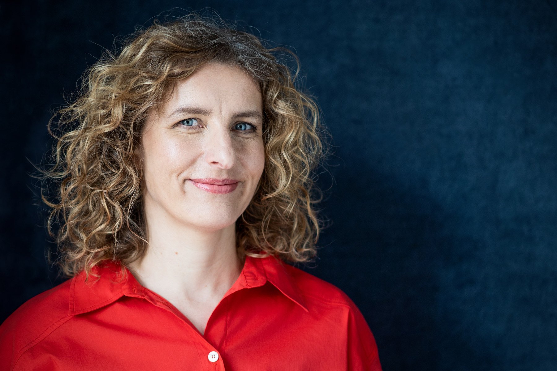 Nahaufnahme einer lächelnden Frau mit blauen Augen und gelocktem, schulterlangem Haar vor dunklem Hintergrund, trägt rotes Hemd.