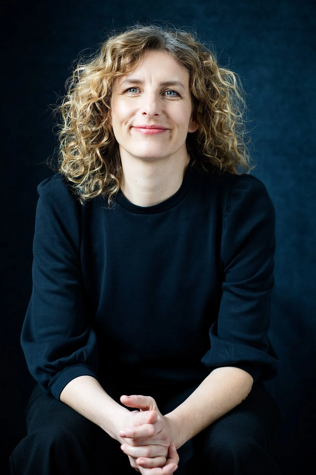 Friederike Sternberg mit lockigem, schulterlangem Haar, die ein schwarzes Oberteil trägt, lächelt vor einem dunklen Hintergrund.