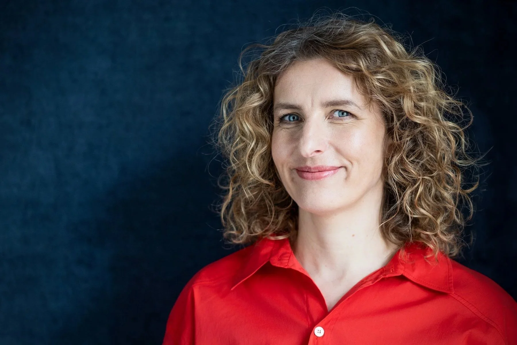 Porträt von Friederike Sternbergmit blonden, lockigen Haaren, trägt eine rote Bluse vor dunklem Hintergrund.