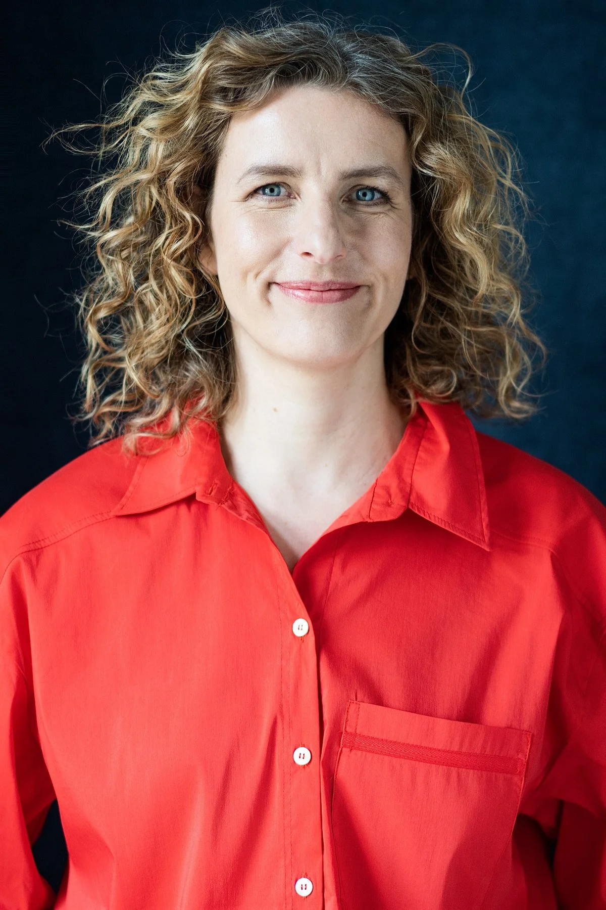 Eine Frau mit lockigem, blonden Haar, trägt eine rote Bluse vor einem dunklen Hintergrund.