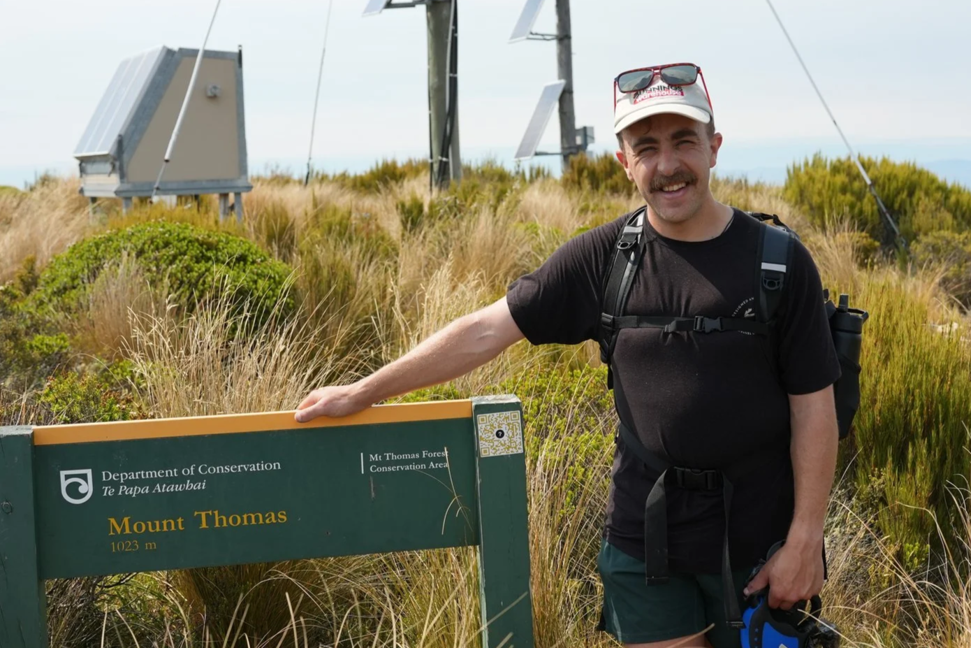A man with a mustache smiling and wearing a black t-shirt, cap, and backpack, standing next to a sign for Mount Thomas at 1023 meters height, in a grassy area with radio towers in the background.
