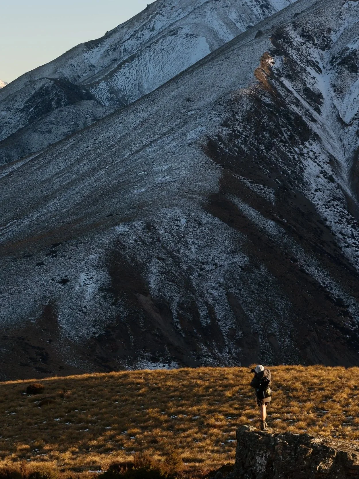 A person with a backpack and a white helmet standing on a grassy area with small patches of snow, overlooking a large mountain with snow patches and rocky terrain in the background during twilight.