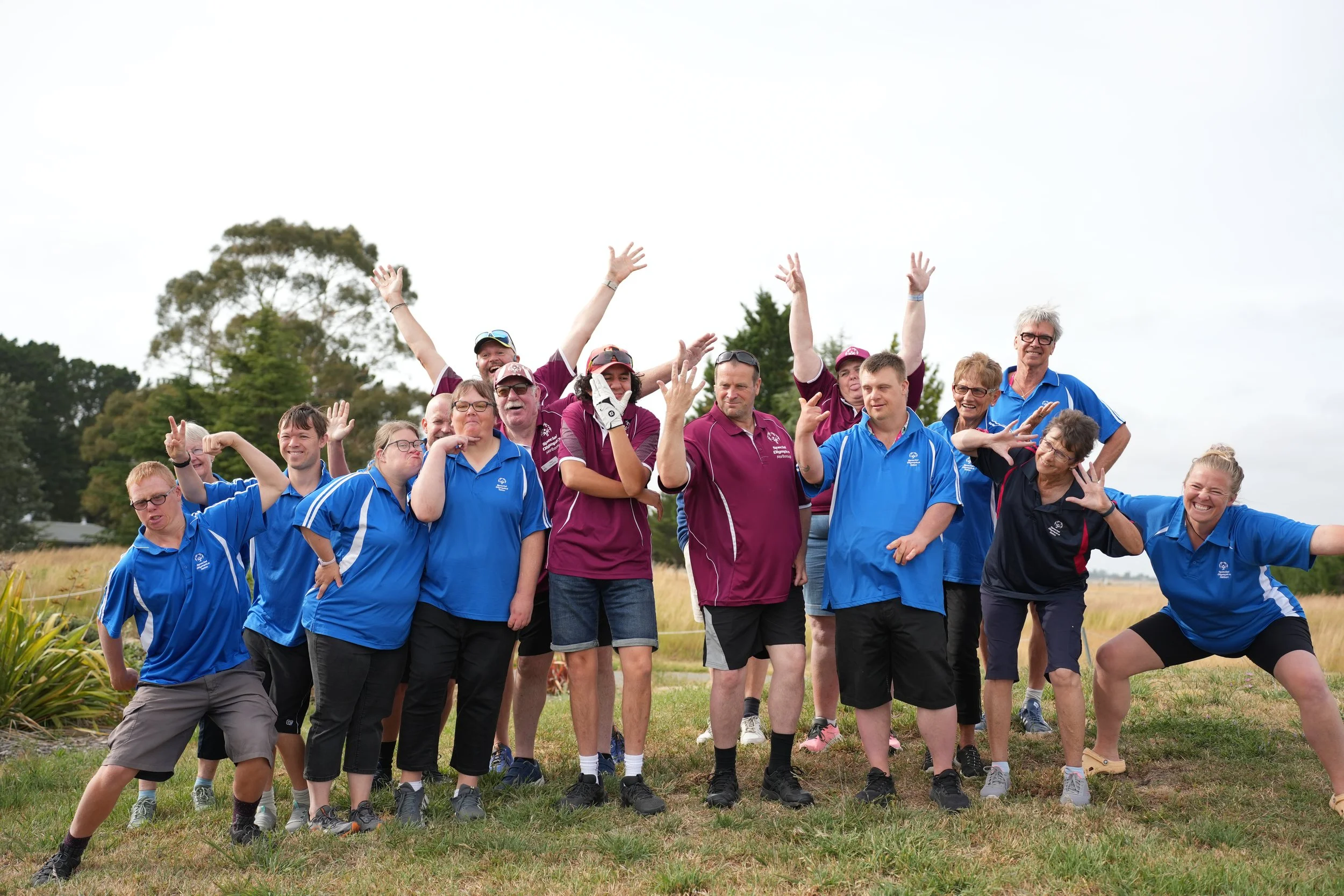 A group of people wearing sporty blue and maroon shirts, posing outdoors in a grassy area with trees and cloudy sky in the background, smiling and making playful gestures.