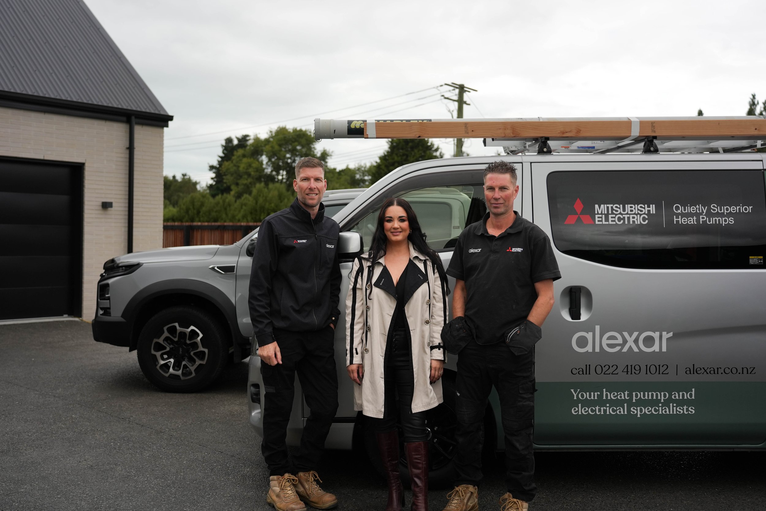 Three professionals standing in front of a silver Mitsubishi electric heat pump service van with the logo and contact information, outdoors on a cloudy day.