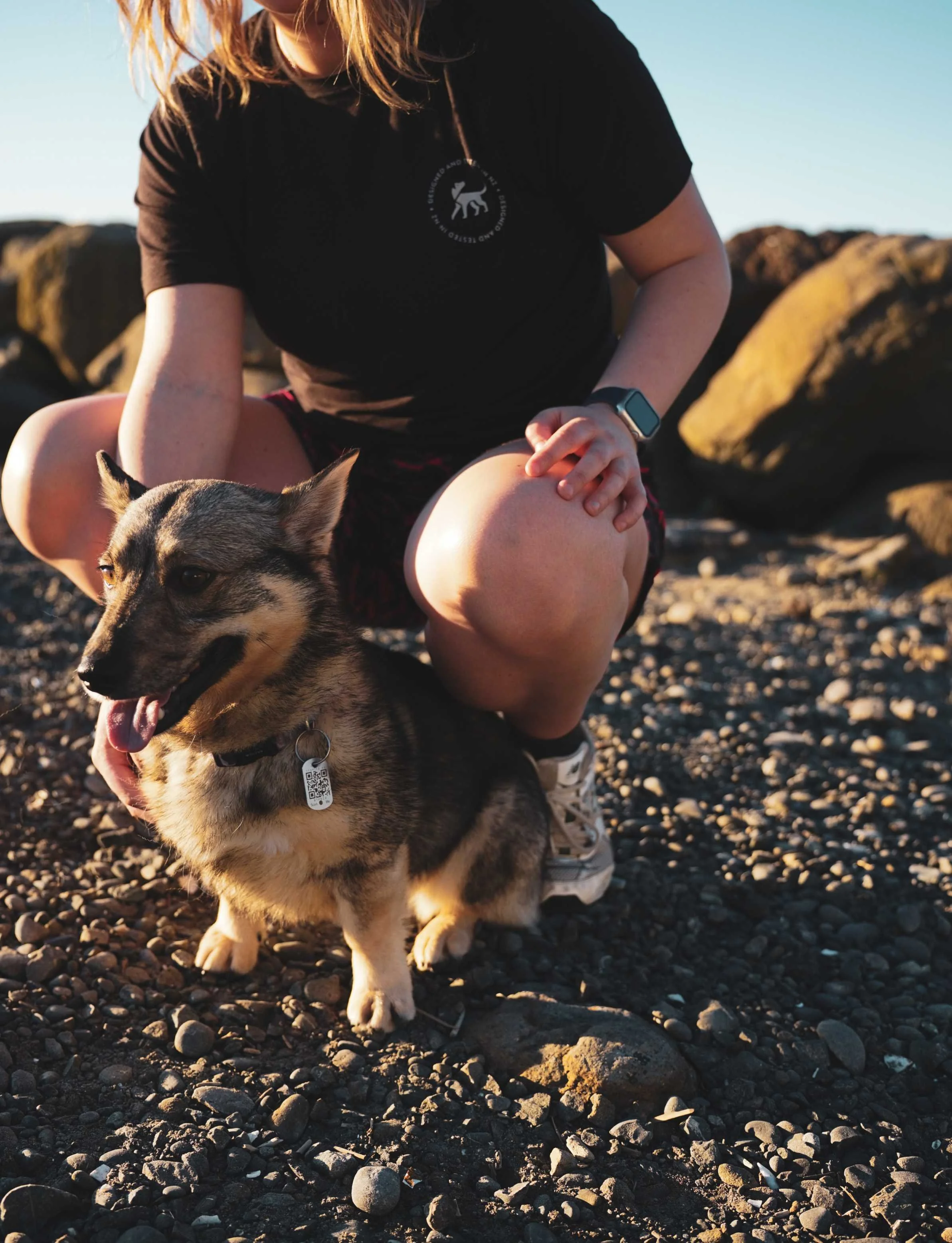 A person with red hair wearing a black t-shirt and shorts crouching on a rocky beach, with a Siberian Husky dog in front of them during sunset.
