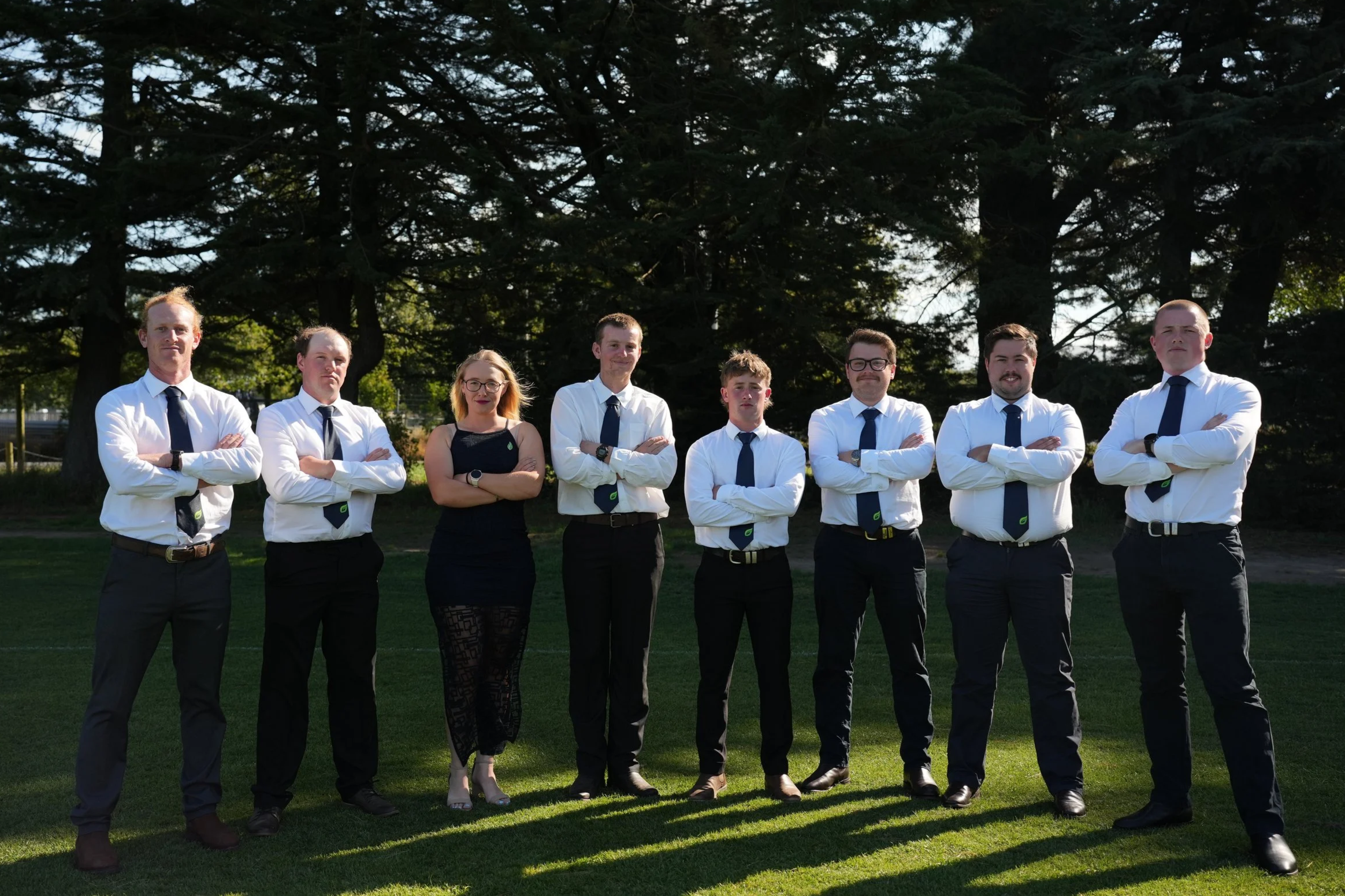 Group of eight people standing outdoors on grass with trees in the background, all dressed in formal or business casual attire with crossed arms.