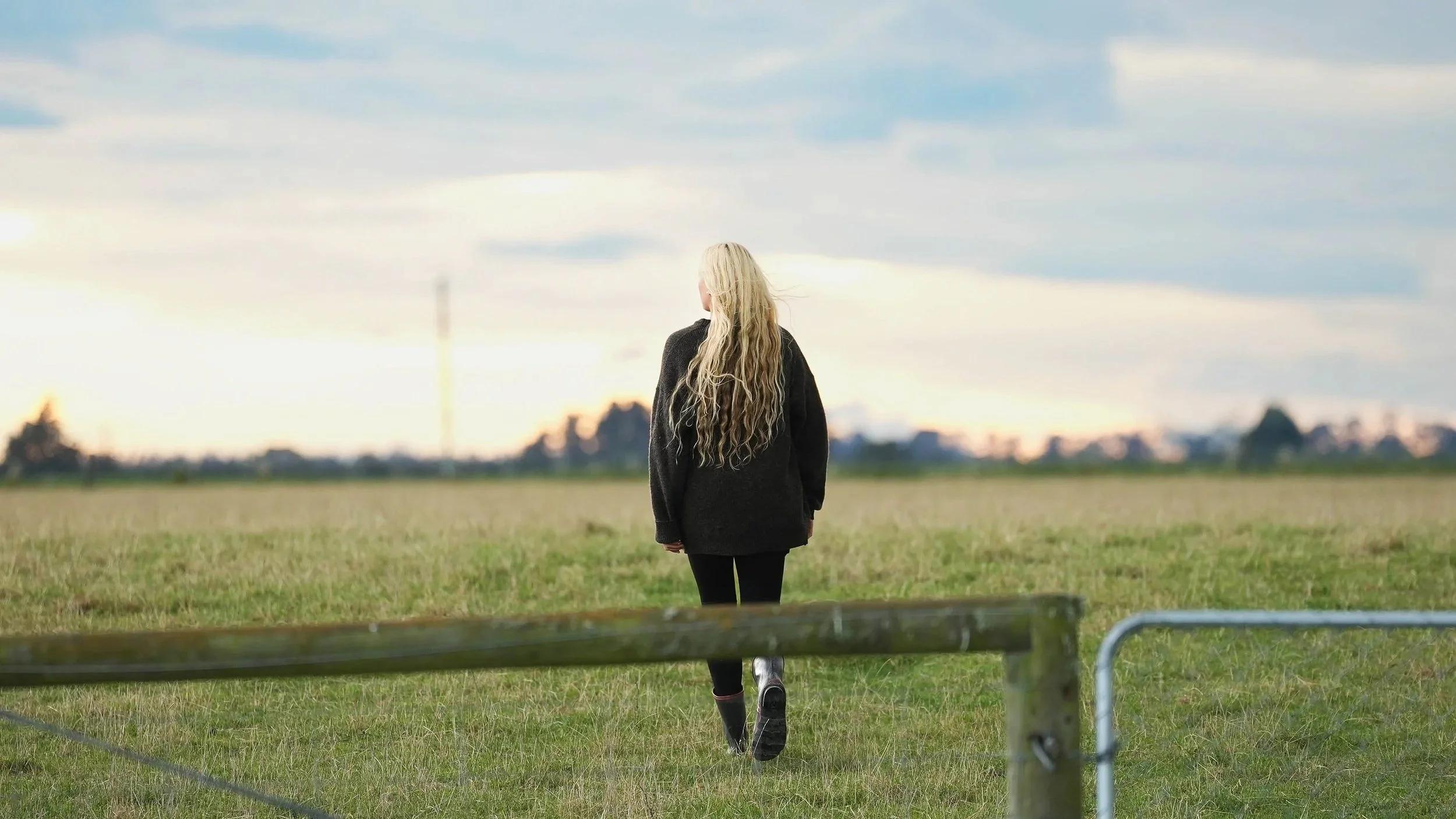 A woman with long blonde hair, wearing a black jacket and black pants, walking in an open grassy field during a cloudy day, with a city skyline in the distance.