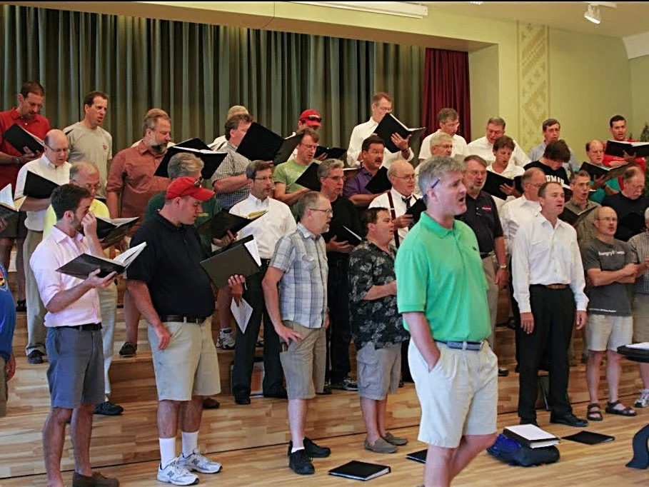 A group of men and women singing in a choir, standing on a stage and holding sheet music, with a green curtain backdrop.