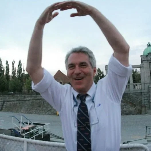 A man with gray hair smiling and holding his hands above his head in a rounded shape outdoors.