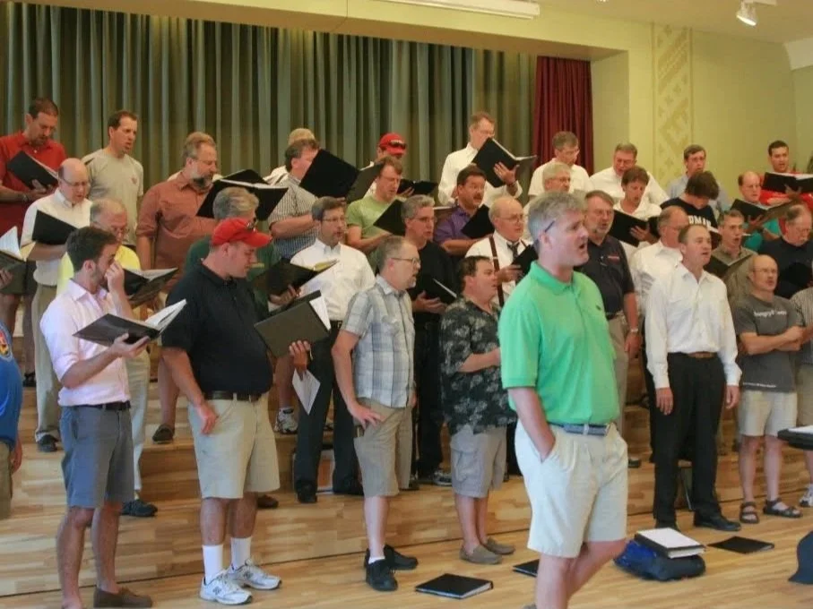 A group of men singing, some holding black folders, in a room with wood flooring and green and red curtains.