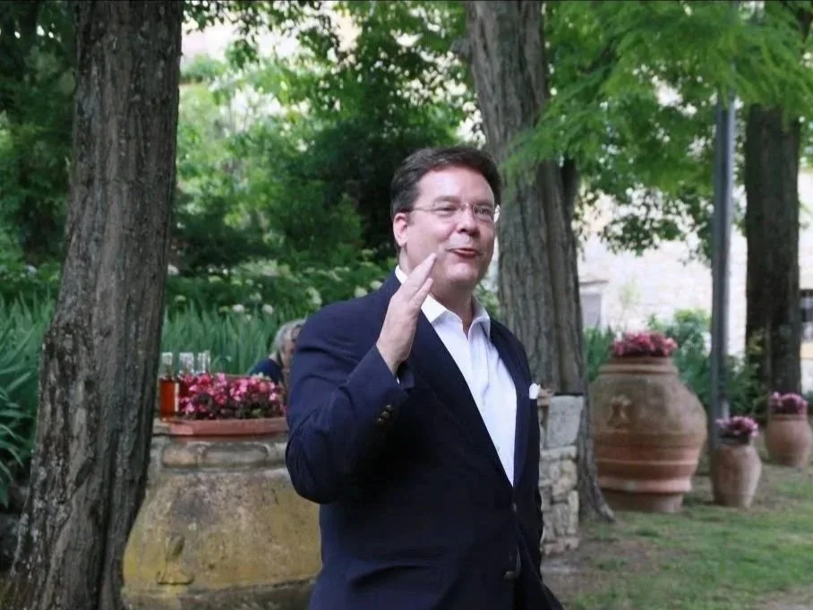 A man in a navy blazer and white shirt, speaking or gesturing outdoors with trees and large potted plants in the background.