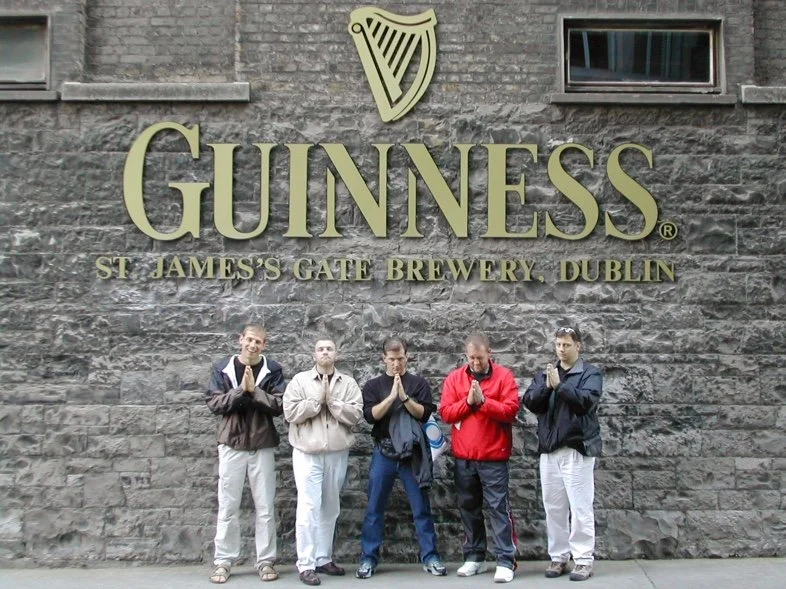 Five Mastersingers standing in a row in front of a large Guinness sign at St. James's Gate Brewery in Dublin as part of the UK Tour in 2003. They are holding their hands in a prayer position.