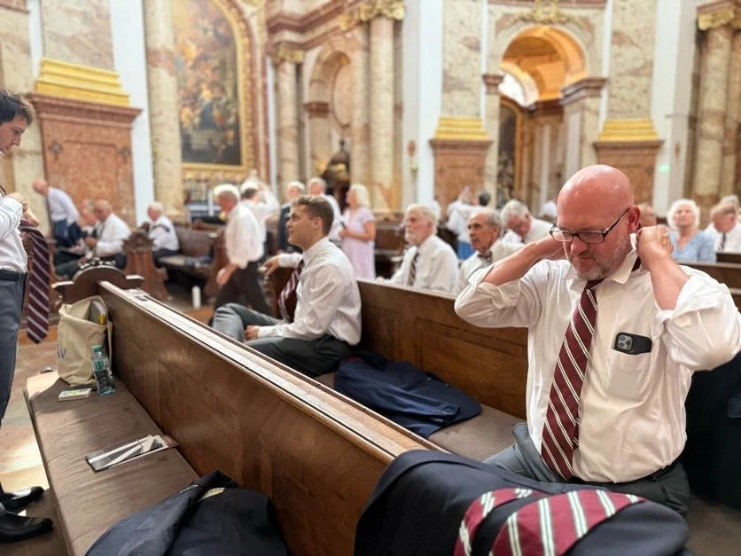 A man in a white shirt and striped tie adjusting his glasses inside a church, with others seated on benches in the background.
