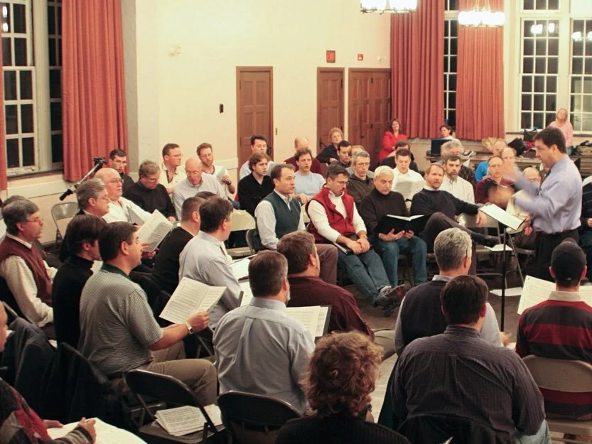 A choir rehearsal or performance indoors with multiple seated singers holding sheet music, a conductor directing from front, large windows, and red curtains.