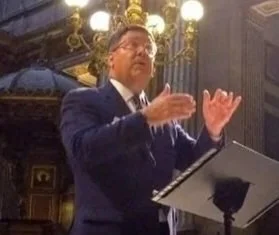 Man in a suit conducting or speaking with hand gestures in an ornate room with chandeliers.