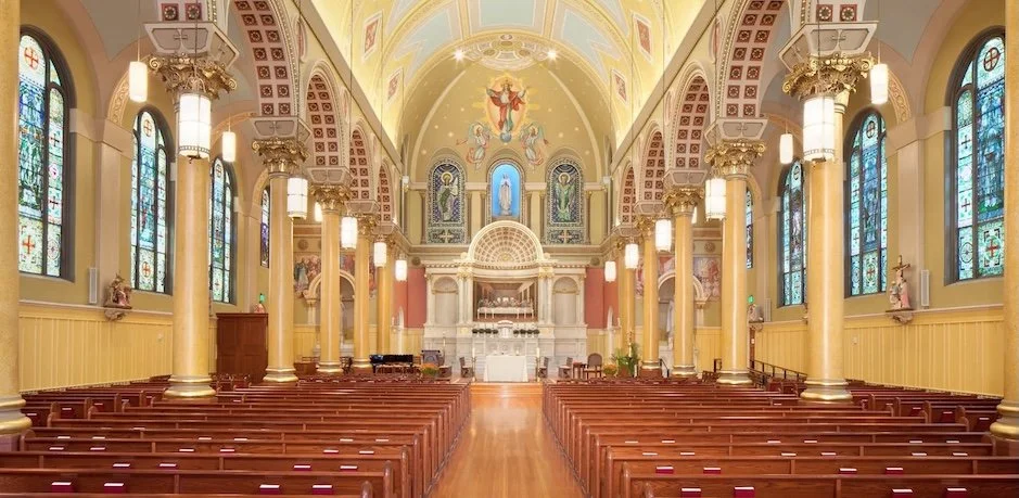 Interior of a church with high arched ceiling, stained glass windows, gold columns, and wooden pews facing an altar.