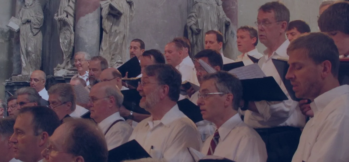Group of men dressed in white shirts, participating in a choir or singing event in a grand hall with classical statues and artwork in the background.