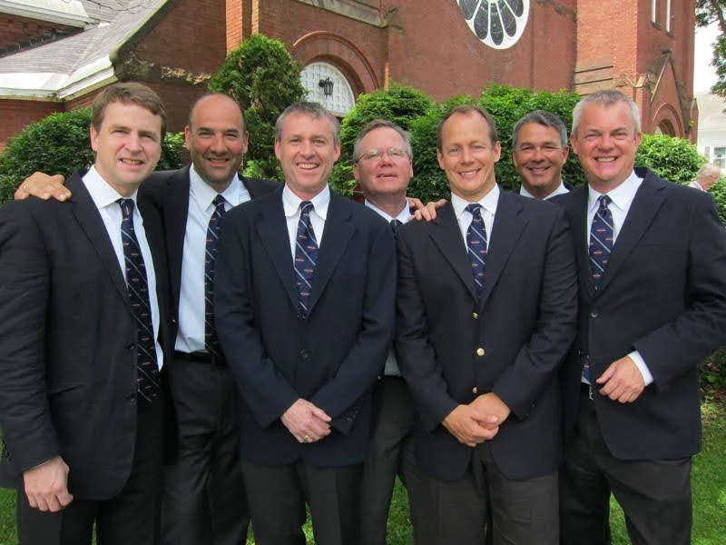 Seven men in suits standing outdoors in front of a brick building with greenery, smiling for a group photo.