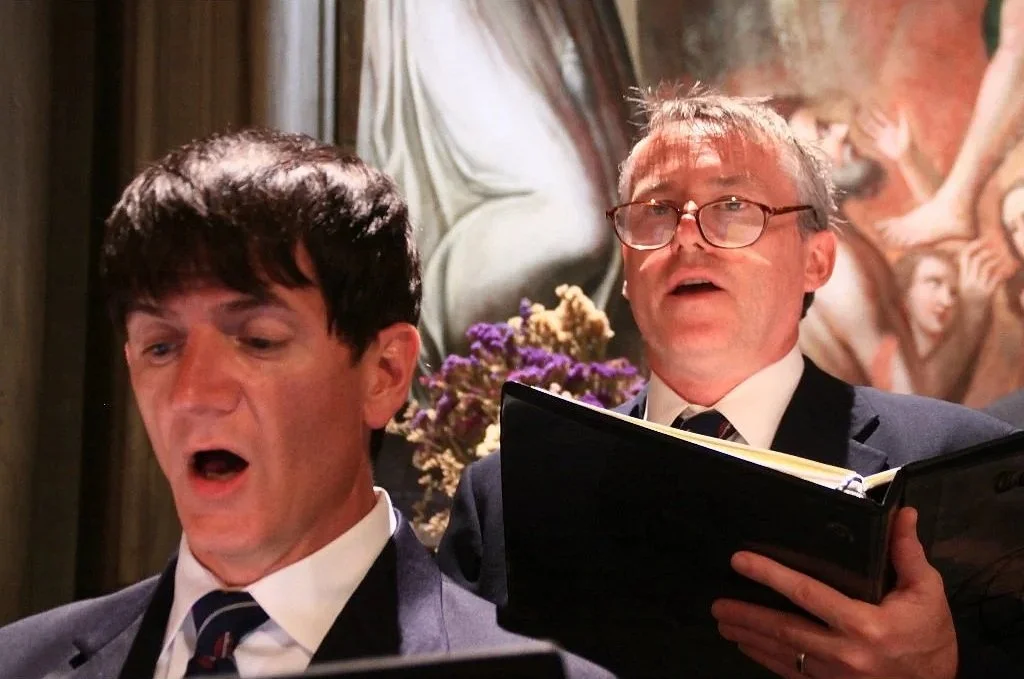 Two Mastersingers dressed in concert attire singing along in a choir, with one holding a music book, in a cathedral.