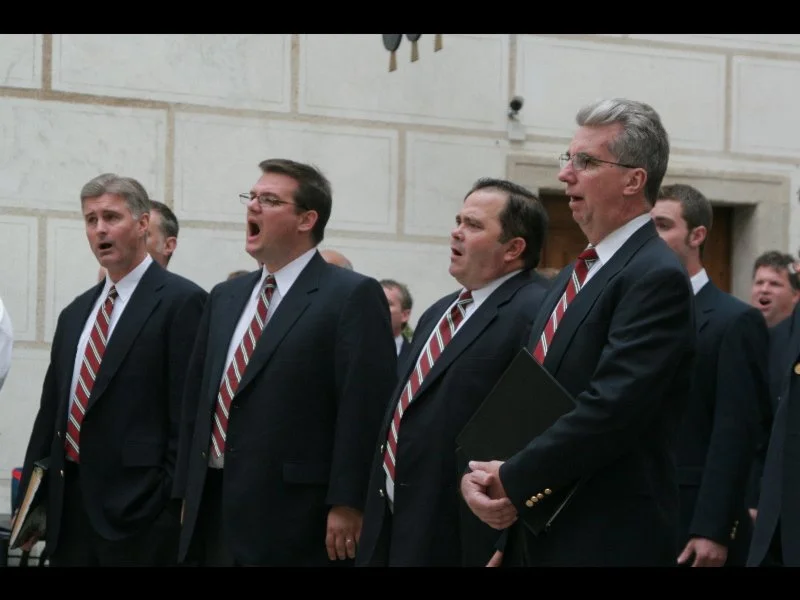 Group of men singing and holding folders, wearing formal suits with striped ties, in a room with white brick walls.