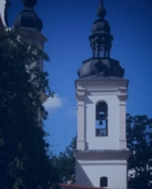 A church steeple with a bell tower, set against a blue sky with trees nearby.