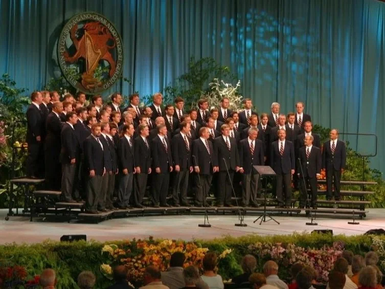 A choir of men and women dressed in dark suits performing on a stage with a blue curtain and greenery backdrop, in front of an audience, with a large emblem featuring a red animal and gold lettering hanging above.