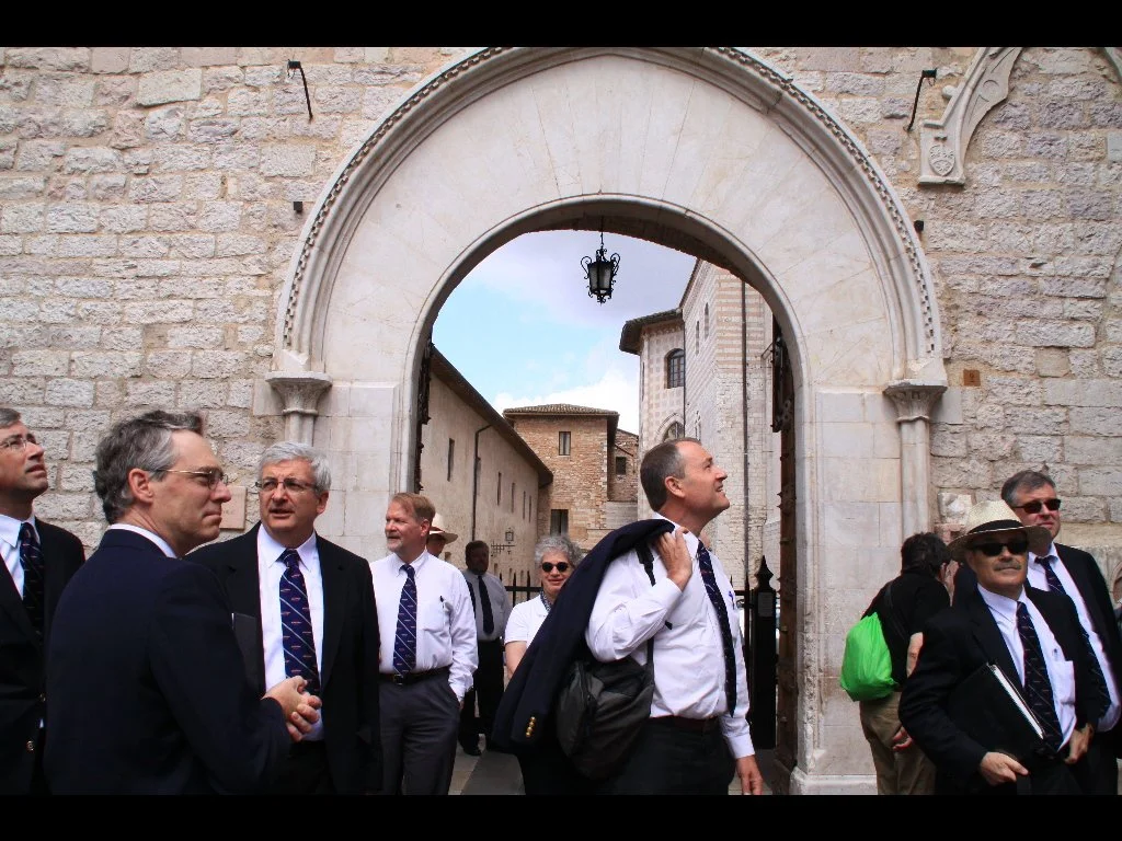 Mastersingers standing outside a historic stone archway, with a view of old brick buildings and a lamppost visible through the arch.