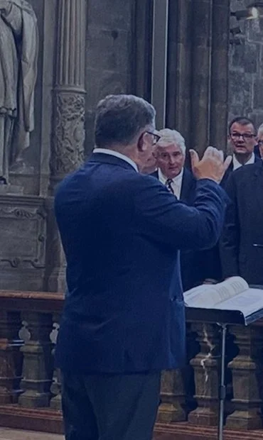 Man in a blue suit conducting a choir or group of singers in a church or cathedral.