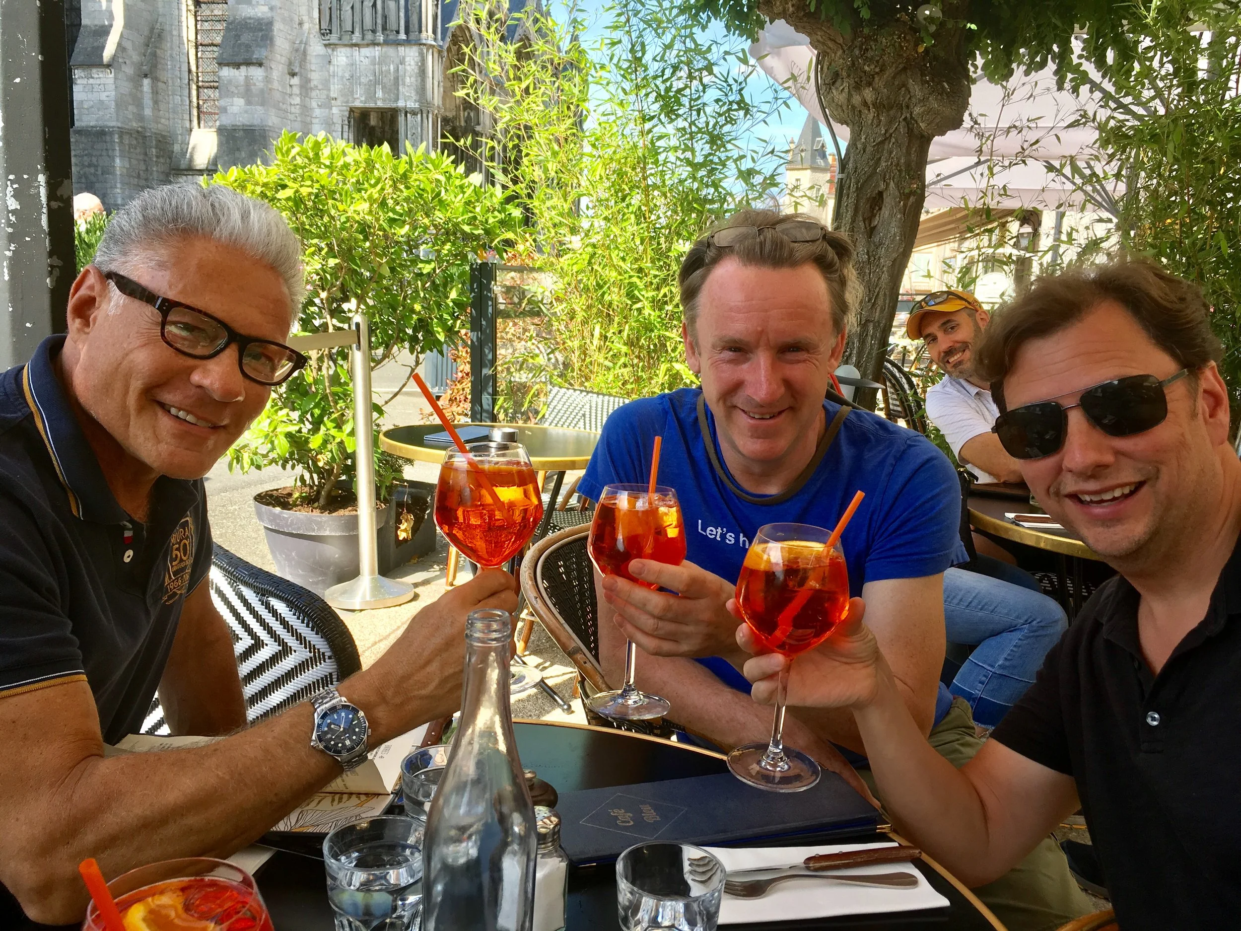 Four men sitting at an outdoor table, smiling and clinking glasses with orange cocktails, surrounded by greenery, with a building in the background.