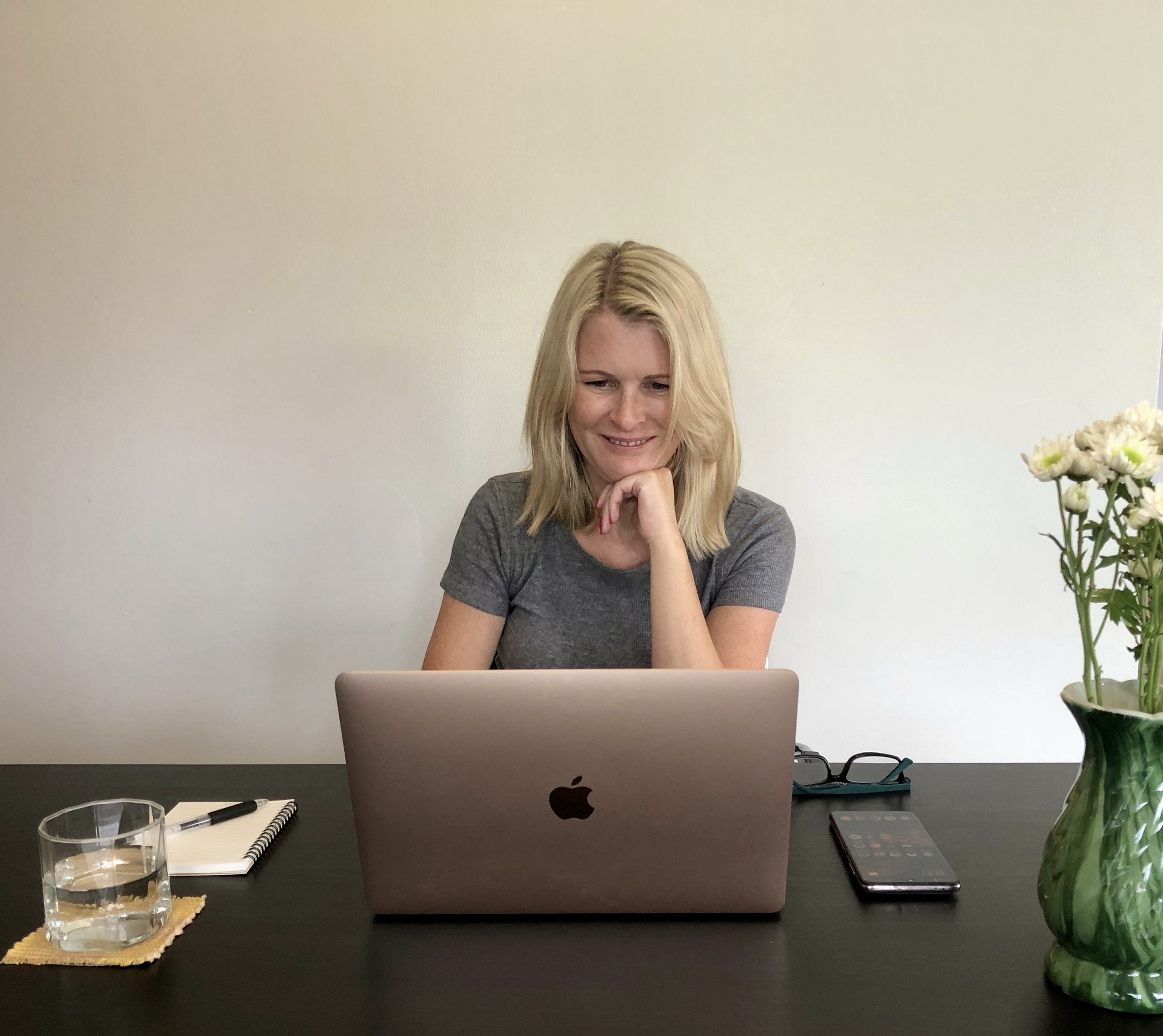 A woman with blonde hair smiling in front of a laptop at a desk with a notebook, pen, glass of water, and a calculator, with a vase of flowers on the right side.