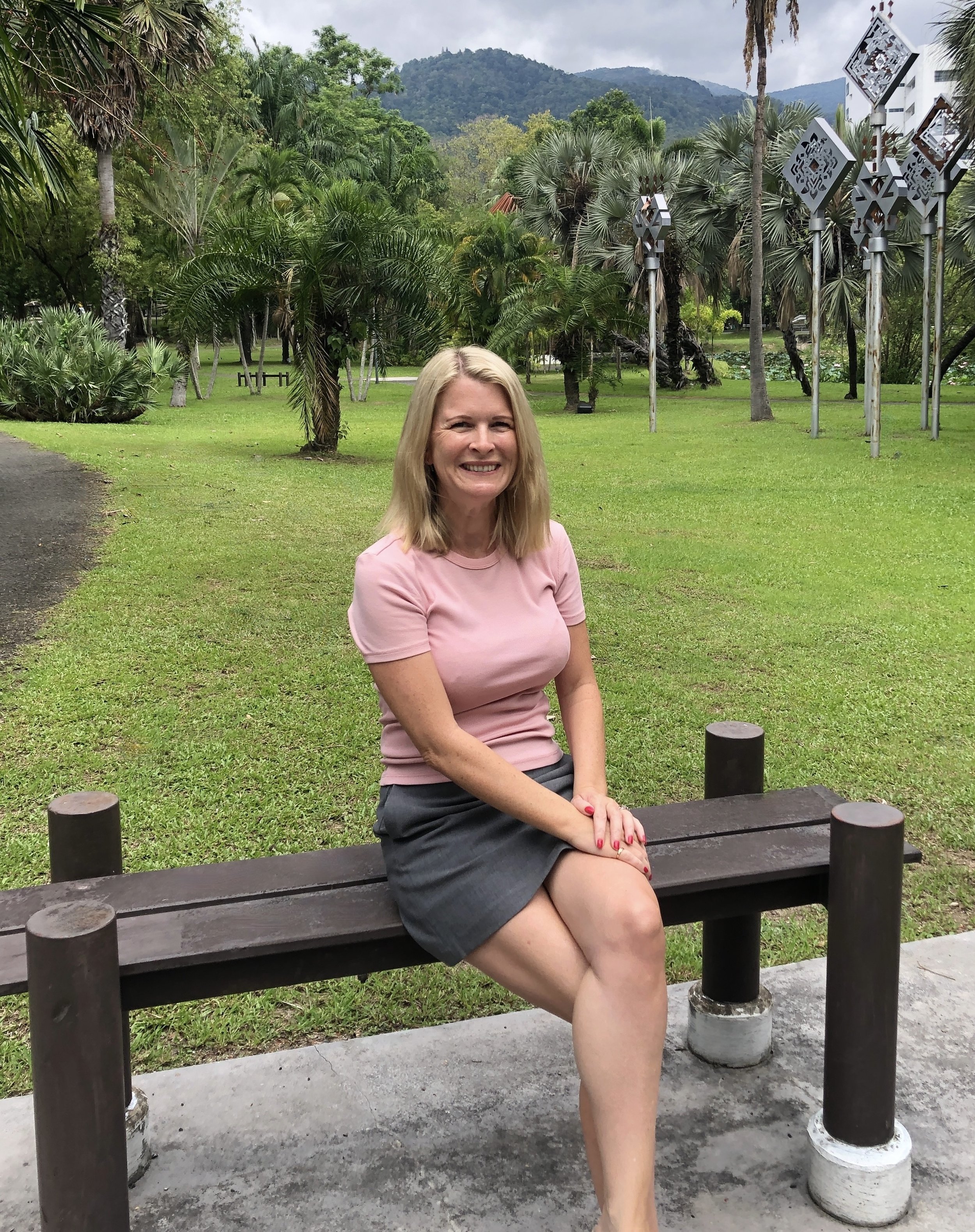 A woman with blonde hair smiling, sitting on a park bench in a lush green outdoor setting with trees and mountains in the background.