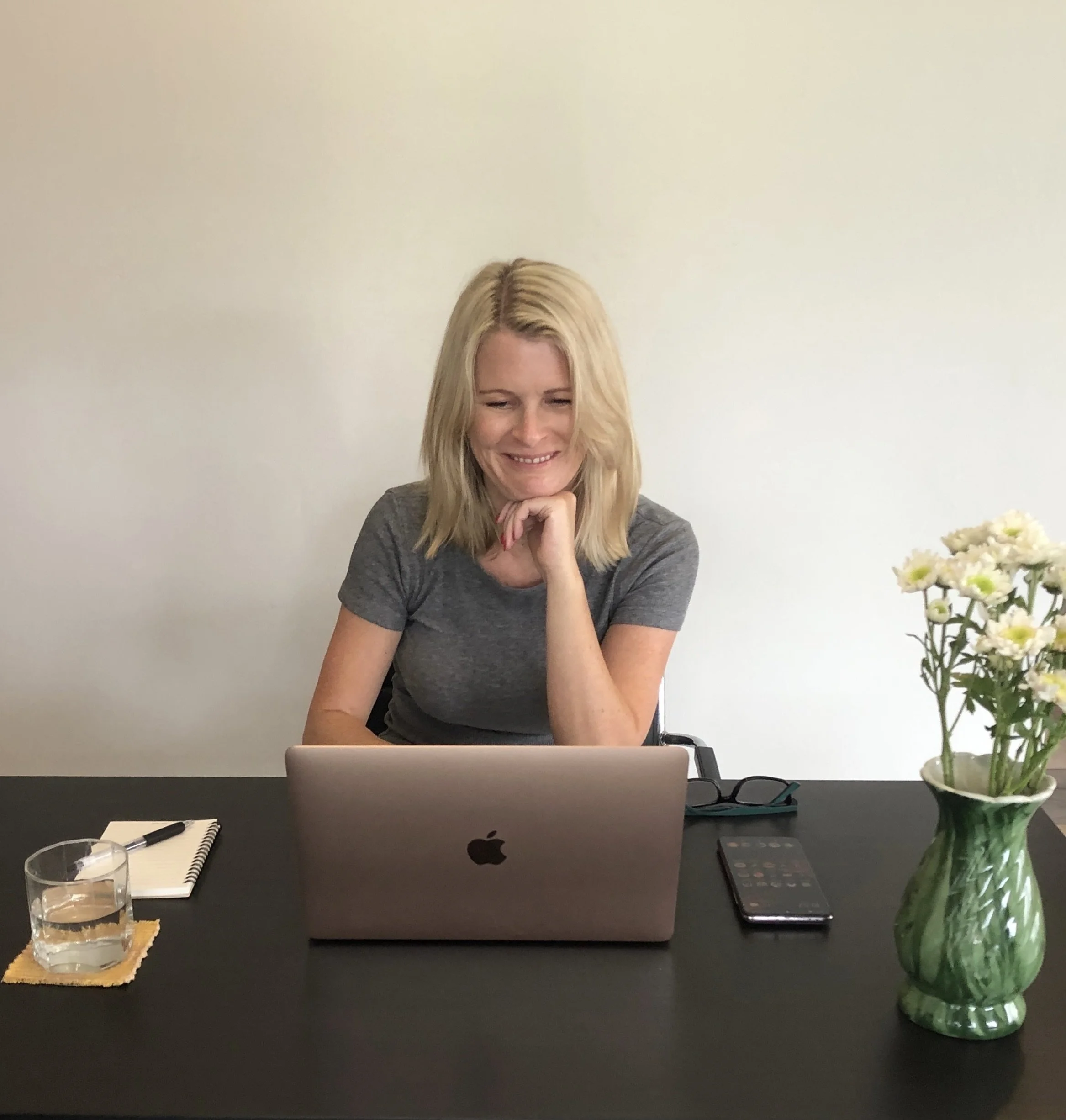 A smiling woman with blonde hair sitting at a black desk with a MacBook, a glass of water, a notebook, a pen, a remote control, and a cell phone. There are white flowers in a vase on the right side of the image, and a plain beige wall in the background.
