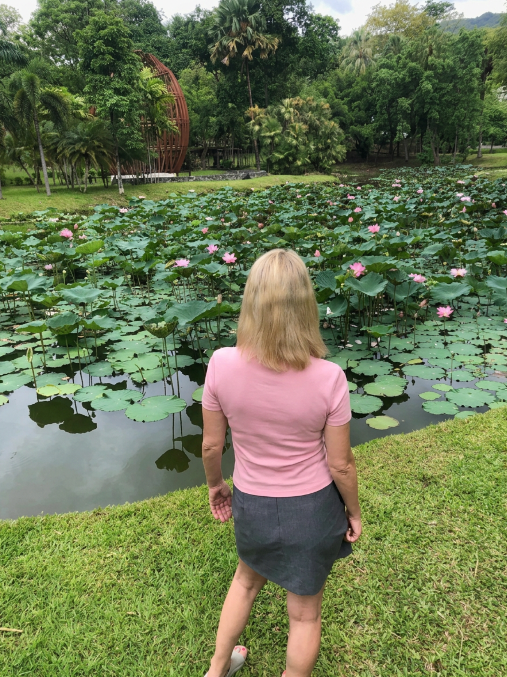 A woman with blonde hair wearing a pink shirt and dark skirt stands by a pond with pink water lilies. She is facing away from the camera, looking at the pond. There are trees and a wooden sculpture in the background.
