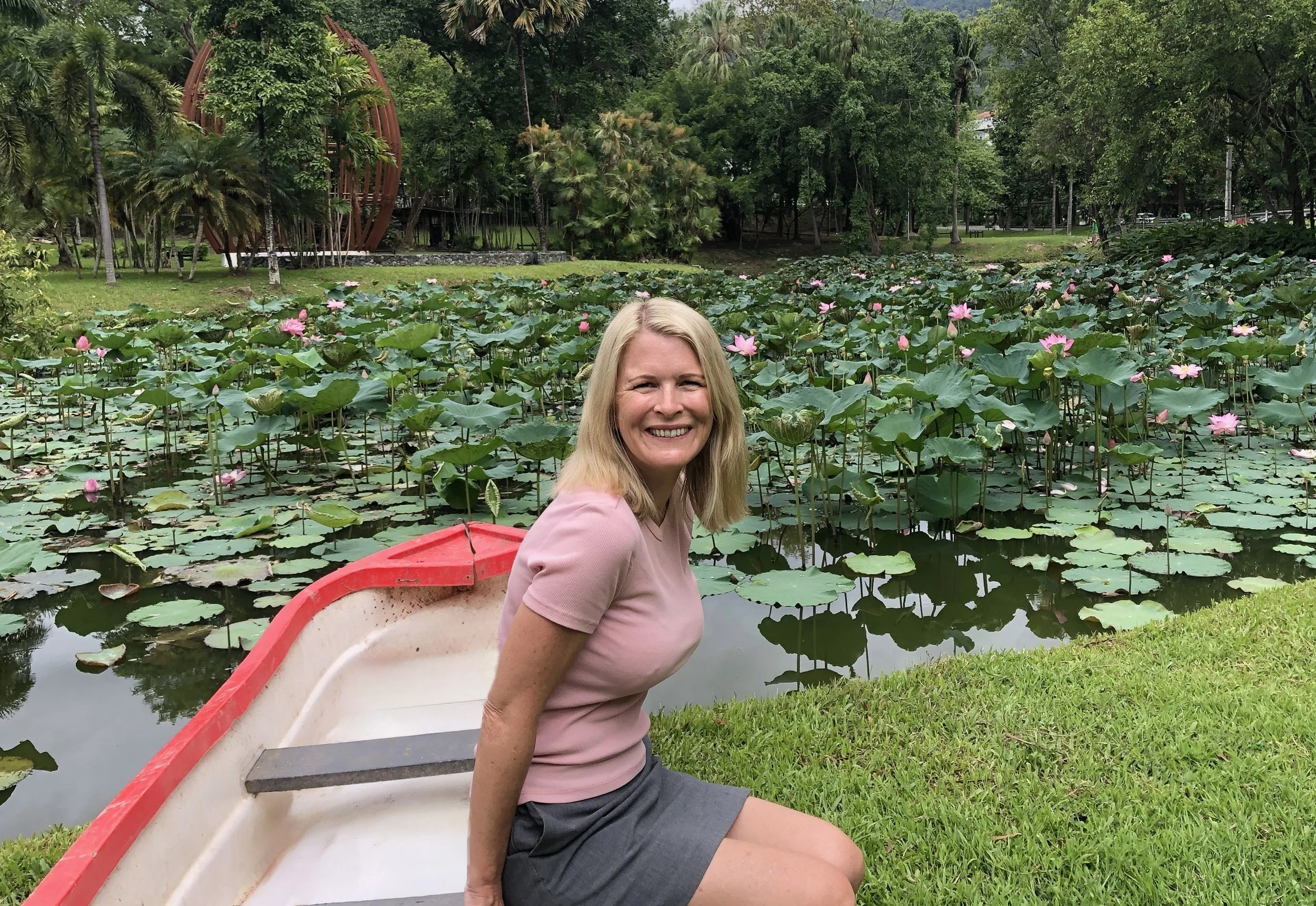Woman with blonde hair smiling while sitting on a boat in a pond filled with pink water lilies, surrounded by lush green trees and plants.